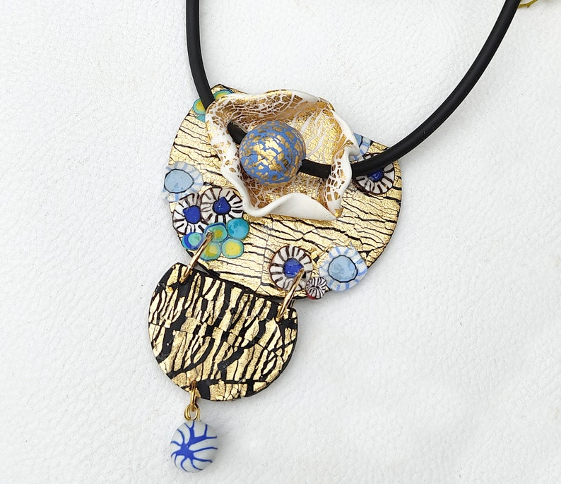 K&ouml;nnte beinhalten: Eine goldene und schwarze Halskette mit einem Anh&auml;nger in Form einer wei&szlig;en Blume mit blauen und goldenen Akzenten. Der Anh&auml;nger ist aus Polymer Clay gefertigt und hat eine strukturierte Oberfl&auml;che.