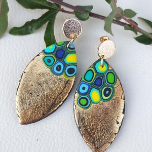 Może przedstawiać: Para złotych kolczyków w kształcie liścia z kolorowym wzorem mozaiki. Mozaika zawiera niebieskie, zielone i żółte okręgi. Kolczyki mają teksturowane złote wykończenie i mały, okrągły złoty sztyft.