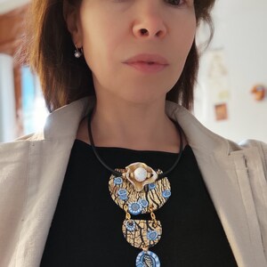 K&ouml;nnte beinhalten: Eine Statement-Halskette mit einem mehrschichtigen Design aus goldenen, wei&szlig;en und blauen Keramikteilen. Die Kette hat ein schwarzes Band und einen gro&szlig;en, runden Anh&auml;nger mit einer craquel&eacute;-Oberfl&auml;che.
