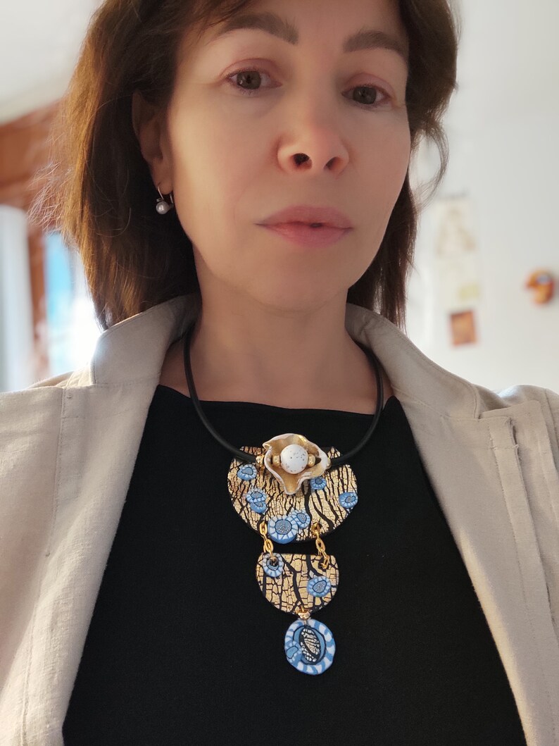 K&ouml;nnte beinhalten: Eine Statement-Halskette mit einem mehrschichtigen Design aus goldenen, wei&szlig;en und blauen Keramikteilen. Die Kette hat ein schwarzes Band und einen gro&szlig;en, runden Anh&auml;nger mit einer craquel&eacute;-Oberfl&auml;che.