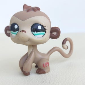 May include: A small, light brown monkey figurine with a large head and oversized eyes. The eyes are black with teal accents and painted eyelashes. The monkey has dark brown ears, hands, and feet, a curled tail, and the red letters LPS on its left leg.