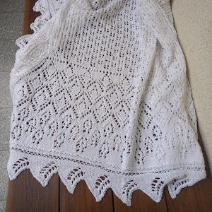 May include: A white, open-weave shawl with a delicate, lacy pattern. The shawl features a scalloped edge with a leaf-like design. The fabric is draped over a wooden surface, showcasing the intricate knitwork and texture of the garment.