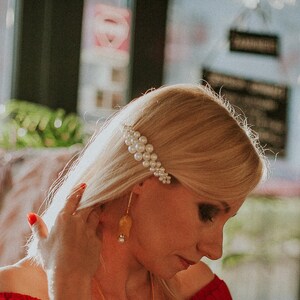 May include: A woman with blonde hair wearing a red off-the-shoulder top and a pearl hair clip. She is wearing gold earrings and a necklace.