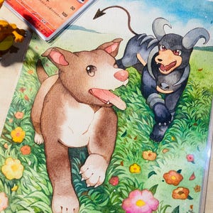 K&ouml;nnte beinhalten: Aquarellmalerei von zwei Hunden in einem Blumenfeld. Einer ist braun, der andere schwarz mit H&ouml;rnern. Eine Pok&eacute;mon-Karte und ein roter Bleistift befinden sich im Hintergrund.