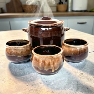 May include: A brown ceramic cooking pot with a lid and two handles, accompanied by three small matching bowls. The bowls have a two-tone design, with a brown base and a lighter, dripped glaze. Steam rises from the pot, suggesting it contains hot food.