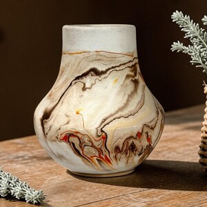 May include: A decorative vase with a unique marbled design in shades of cream, brown, and red. The vase has a wide base and a narrower neck, with a light-colored rim. The vase is sitting on a wooden surface.