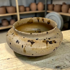 May include: A low-profile ceramic bowl with a wide, rounded body and a short, upright rim. The exterior features a light beige, speckled glaze with dark brown and black splatters distributed across the surface, creating a textured appearance.