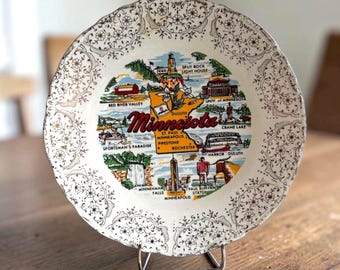 May include: A decorative ceramic plate with a scalloped edge and a brown floral border. The center features a colorful map of Minnesota with illustrations of landmarks like Split Rock Lighthouse, Minnehaha Falls, and the Paul Bunyan statue.