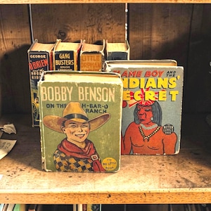 May include: A collection of vintage Big Little Books on a wooden shelf. The books feature colorful covers with titles like "Bobby Benson on the H-Bar-O Ranch" and "Flame Boy and the Indians Secret". The books are in various states of wear.