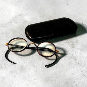 May include: Round eyeglasses with a dark brown frame and black temples, paired with a black leather eyeglass case. The glasses have a gold-colored accent at the hinges. The lenses are clear.
