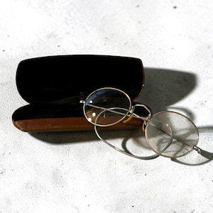 May include: Vintage round eyeglasses with a gold-colored frame and clear lenses. The glasses are next to an open, brown leather case with a black interior. The case is slightly open, and the glasses are resting on a light-colored surface.