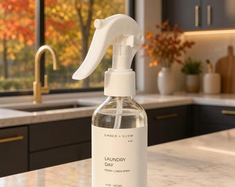 May include: A clear glass spray bottle with a white trigger nozzle and a white label. The label reads Ember + Glow, Laundry Day, Room + Linen Spray, 4 oz, Vegan, Fleming Island, FL. The bottle contains a transparent liquid and has a white cap.