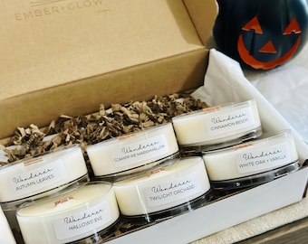 May include: A cardboard gift box containing six small round candles with white wax and wooden wicks. Each candle has a label reading Wanderer followed by scent names: Autumn Leaves, Campfire Marshmallow, Cinnamon Besom, Twilight Orchard, Hallows' Eve, and White Oak + Vanilla. The box lid is printed with Ember + Glow.
