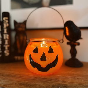 May include: A round orange glass candle holder shaped like a jack-o'-lantern with a black painted face featuring triangular eyes and a wide grinning mouth. The container includes a thin dark metal wire handle attached to the top rim.