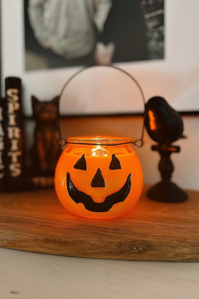 May include: A round orange glass candle holder shaped like a jack-o'-lantern with a black painted face featuring triangular eyes and a wide grinning mouth. The container includes a thin dark metal wire handle attached to the top rim.