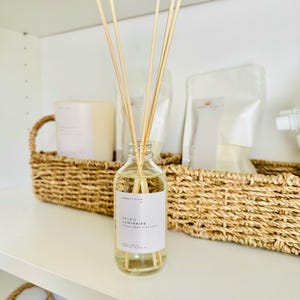 May include: A clear glass reed diffuser bottle filled with light yellow liquid and several wooden reeds. The white label features the text Ember + Glow, Soleil Luminaire, Clean Reed Diffuser, 4 oz, Vegan, Fernandina Island, FL.