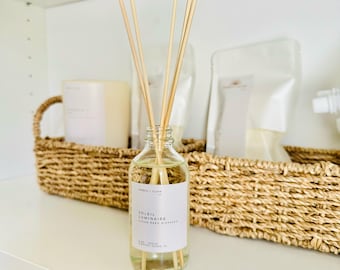 Op de afbeelding: Een doorzichtige glazen rietdiffuserfles gevuld met lichtgele vloeistof en verschillende houten rietjes. Het witte etiket bevat de tekst Ember + Glow, Soleil Luminaire, Clean Reed Diffuser, 4 oz, Vegan, Fernandina Island, FL.