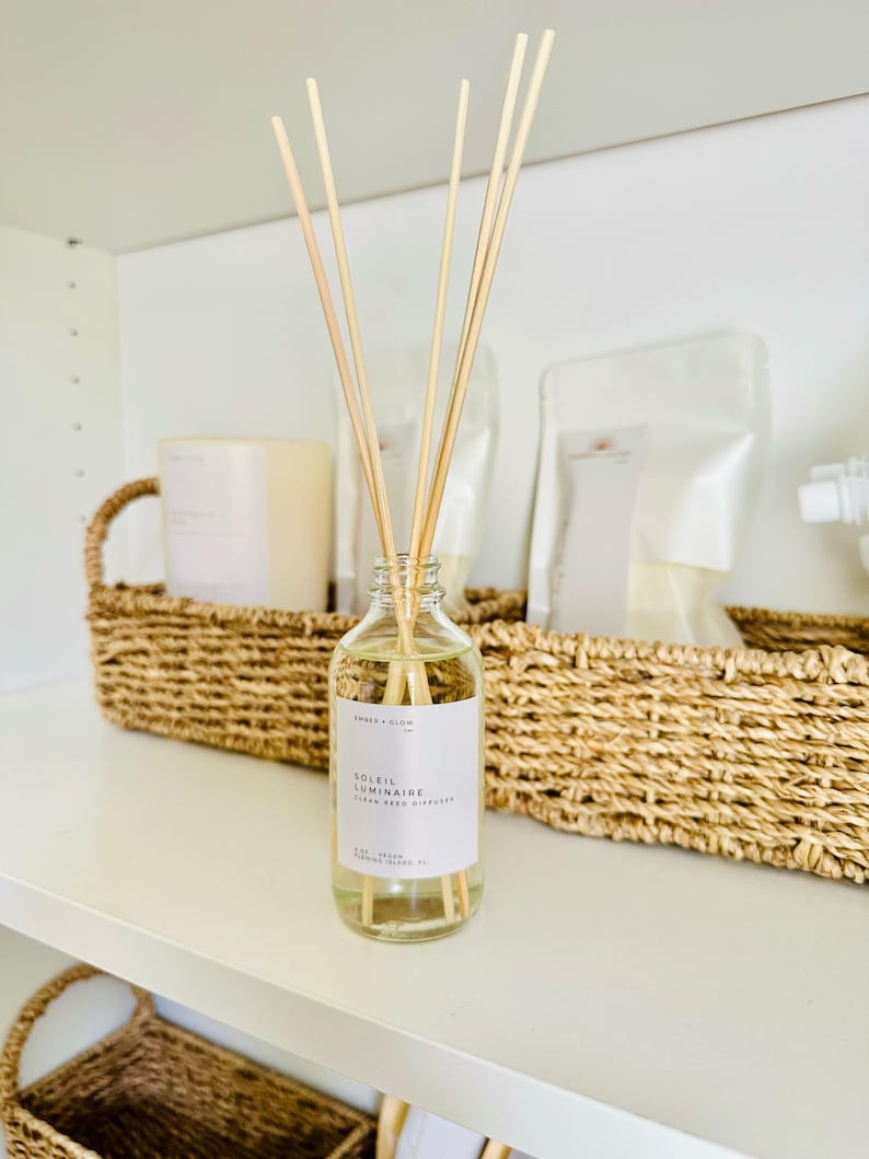 May include: A clear glass reed diffuser bottle filled with light yellow liquid and several wooden reeds. The white label features the text Ember + Glow, Soleil Luminaire, Clean Reed Diffuser, 4 oz, Vegan, Fernandina Island, FL.