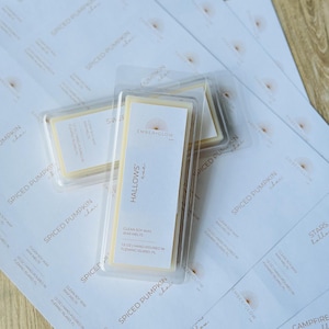 May include: Two clear plastic clamshell containers holding rectangular off-white soy wax melts. The labels feature the text Ember + Glow, Hallows' Eve, and 1.5 oz | Hand-poured in Fleming Island, FL. They rest on a sheet of printed product labels.