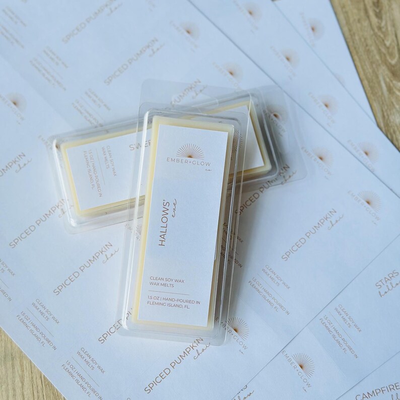 May include: Two clear plastic clamshell containers holding rectangular off-white soy wax melts. The labels feature the text Ember + Glow, Hallows' Eve, and 1.5 oz | Hand-poured in Fleming Island, FL. They rest on a sheet of printed product labels.