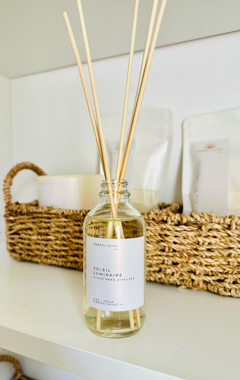 May include: A clear glass reed diffuser bottle containing light-colored liquid and four wooden reeds. The white label reads EMBER + GLOW, SOLEIL LUMINAIRE, CLEAN REED DIFFUSER, 4 OZ, VEGAN, FLEMING ISLAND, FL in black text.