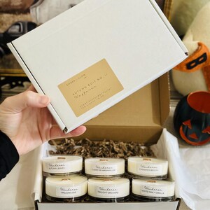 Op de afbeelding: Een witte kartonnen doos met zes kleine ronde kaarsen met houten lonten. Het deksel heeft een beige label met de tekst Ember + Glow, Autumn Edit No. 1, Wanderer, Scent Discovery Flight, Tea Lights 1.9 oz. De kaarsen zijn gelabeld met Autumn Leaves, Campfire Marshmallow, Cinnamon Beech, Hallows Eve, Twilight Orchard en White Oak + Vanilla.