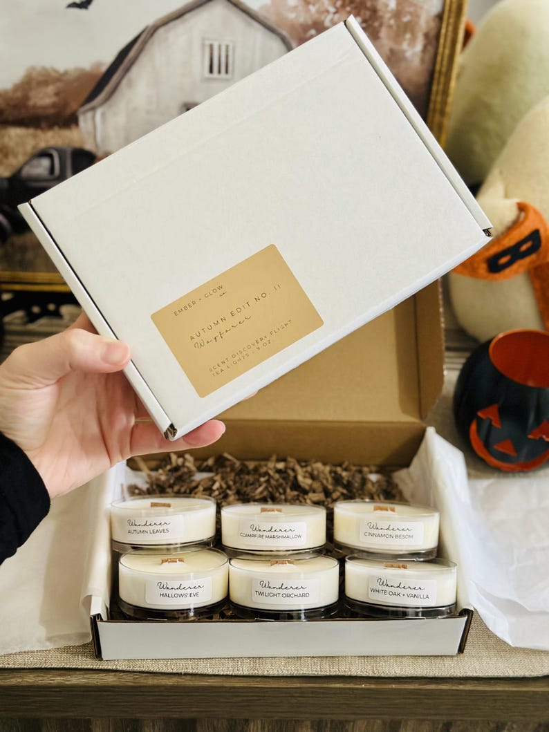 Op de afbeelding: Een witte kartonnen doos met zes kleine ronde kaarsen met houten lonten. Het deksel heeft een beige label met de tekst Ember + Glow, Autumn Edit No. 1, Wanderer, Scent Discovery Flight, Tea Lights 1.9 oz. De kaarsen zijn gelabeld met Autumn Leaves, Campfire Marshmallow, Cinnamon Beech, Hallows Eve, Twilight Orchard en White Oak + Vanilla.