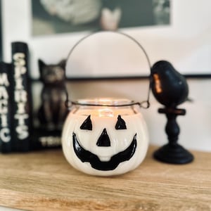 May include: A white glass pumpkin-shaped candle holder with a black painted jack-o'-lantern face featuring triangular eyes and a wide grin. The container has a thin black metal wire handle attached to the rim and contains a lit candle inside.