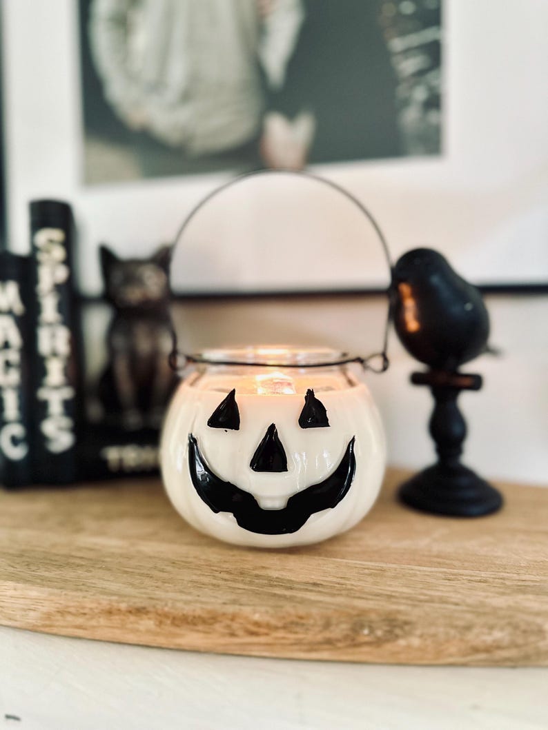 May include: A white glass pumpkin-shaped candle holder with a black painted jack-o'-lantern face featuring triangular eyes and a wide grin. The container has a thin black metal wire handle attached to the rim and contains a lit candle inside.