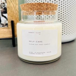 May include: A glass candle jar with a cork lid. The label reads "EMBER GLOW co. SELF-CARE CLEAN SOY WAX CANDLE 16 OZ. 75 HOUR BURN - VEGAN FLEMING ISLAND, FL."
