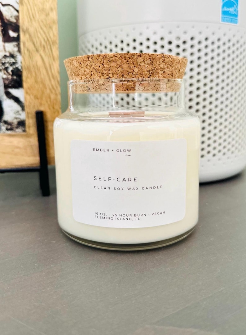 May include: A glass candle jar with a cork lid. The label reads "EMBER GLOW co. SELF-CARE CLEAN SOY WAX CANDLE 16 OZ. 75 HOUR BURN - VEGAN FLEMING ISLAND, FL."