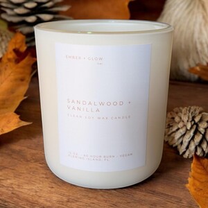 May include: A white frosted glass candle jar with a white label. The label reads "EMBER GLOW" in a small font at the top, followed by "SANDALWOOD + VANILLA" in a larger font, and "CLEAN SOY WAX CANDLE" in a smaller font. The bottom of the label reads "12 OZ 60 HOUR BURN VEGAN FLEMING ISLAND, FL".