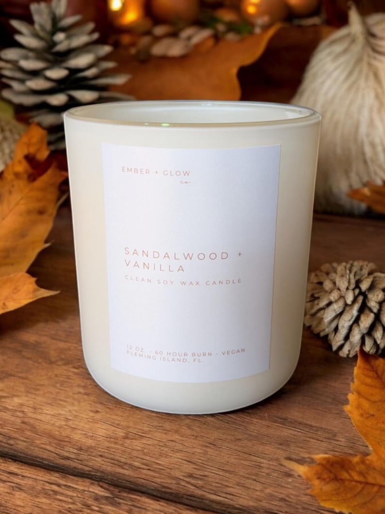 May include: A white frosted glass candle jar with a white label. The label reads "EMBER GLOW" in a small font at the top, followed by "SANDALWOOD + VANILLA" in a larger font, and "CLEAN SOY WAX CANDLE" in a smaller font. The bottom of the label reads "12 OZ 60 HOUR BURN VEGAN FLEMING ISLAND, FL".