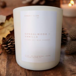 May include: White candle with a white label that reads "EMBER + GLOW" and "SANDALWOOD + VANILLA" in a brown font. The label also says "CLEAN SOY WAX CANDLE", "12 OZ - 60 HOUR BURN - VEGAN", and "FLEMING ISLAND, FL".