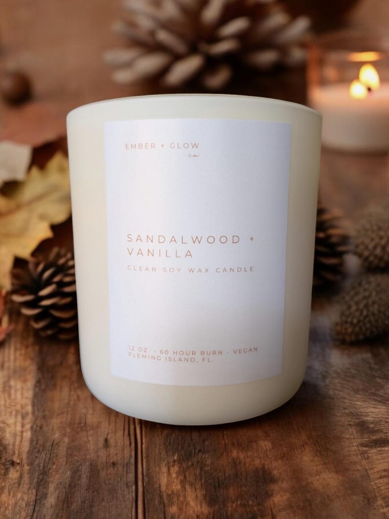 May include: White candle with a white label that reads "EMBER + GLOW" and "SANDALWOOD + VANILLA" in a brown font. The label also says "CLEAN SOY WAX CANDLE", "12 OZ - 60 HOUR BURN - VEGAN", and "FLEMING ISLAND, FL".