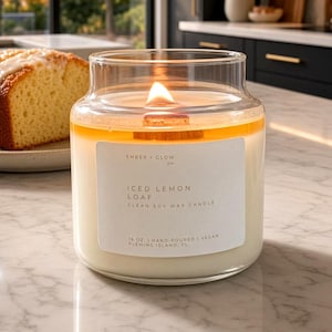May include: A clear glass jar candle with a lit wooden wick and off-white soy wax. The white label reads Ember Glow Co., Iced Lemon Loaf, Clean Soy Wax Candle, 16 oz, Hand-Poured, Vegan, Fleming Island, FL. The candle has a visible melted wax pool.