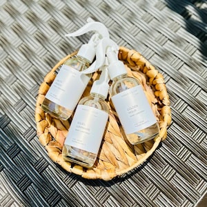 May include: Three clear glass spray bottles with white labels and spray nozzles are arranged in a woven basket. The labels read "Salty Beach", "Soleil Luminaire", and "Golden Hour". The basket sits on a textured, dark surface.