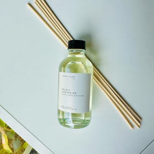 May include: A clear glass bottle filled with light yellow liquid, topped with a black cap, and labeled Ember + Glow Soleil Luminaire Clean Reed Diffuser. A bundle of light-colored wooden reeds is positioned diagonally behind the bottle.