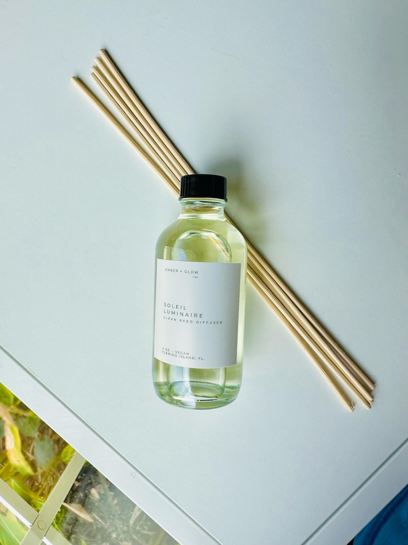 May include: A clear glass bottle filled with light yellow liquid, topped with a black cap, and labeled Ember + Glow Soleil Luminaire Clean Reed Diffuser. A bundle of light-colored wooden reeds is positioned diagonally behind the bottle.