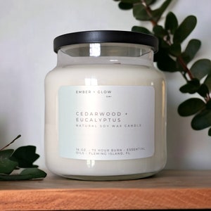May include: A clear glass jar candle with a black lid. The label reads "EMBER + GLOW Co." and "CEDARWOOD + EUCALYPTUS NATURAL SOY WAX CANDLE". The label also states "16 OZ. 75 HOUR BURN - ESSENTIAL OILS - FLEMING ISLAND, FL."