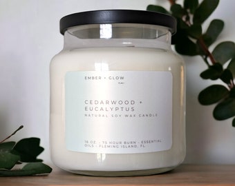 May include: A clear glass jar candle with a black lid. The label reads "EMBER + GLOW Co." and "CEDARWOOD + EUCALYPTUS NATURAL SOY WAX CANDLE". The label also states "16 OZ. 75 HOUR BURN - ESSENTIAL OILS - FLEMING ISLAND, FL."