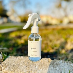 May include: A clear glass spray bottle with a white trigger nozzle and a white label. The label features the text Ember + Glow, Laundry Day, Room + Linen Spray, 4 oz, Vegan, Fleming Island, FL in black font against a white background.