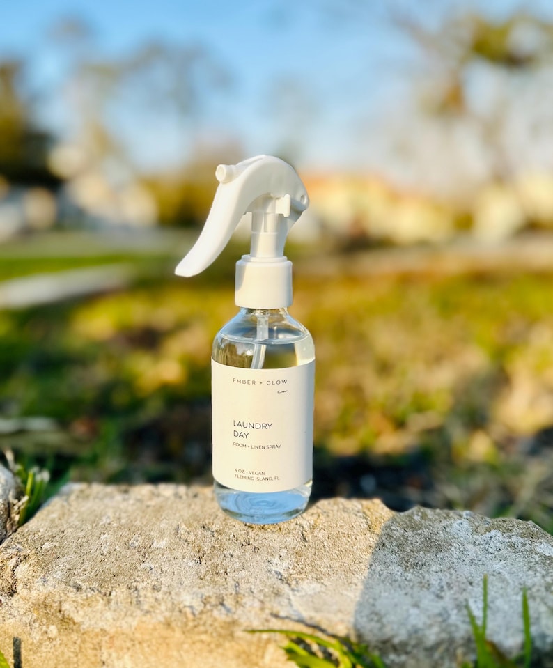 May include: A clear glass spray bottle with a white trigger nozzle and a white label. The label features the text Ember + Glow, Laundry Day, Room + Linen Spray, 4 oz, Vegan, Fleming Island, FL in black font against a white background.