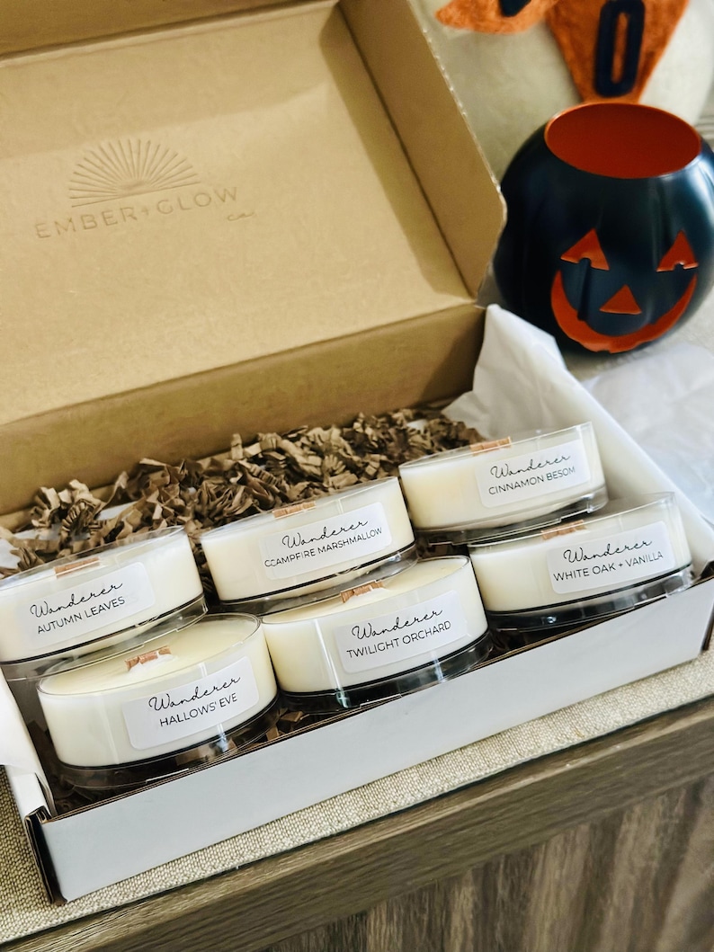 Op de afbeelding: Een kartonnen geschenkdoos met zes kleine ronde kaarsen met witte was en houten lonten. Elke kaars heeft een label met de tekst Wanderer gevolgd door de geurnamen: Autumn Leaves, Campfire Marshmallow, Cinnamon Besom, Twilight Orchard, Hallows' Eve en White Oak + Vanilla. Het deksel is bedrukt met Ember + Glow.