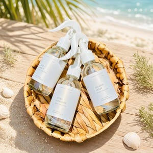 May include: Three clear glass spray bottles with white trigger nozzles and white labels, each containing a liquid. The labels feature the text Ember & Glow, with individual names Salty Beach, Soleil Luminaire, and Golden Hour printed on them.