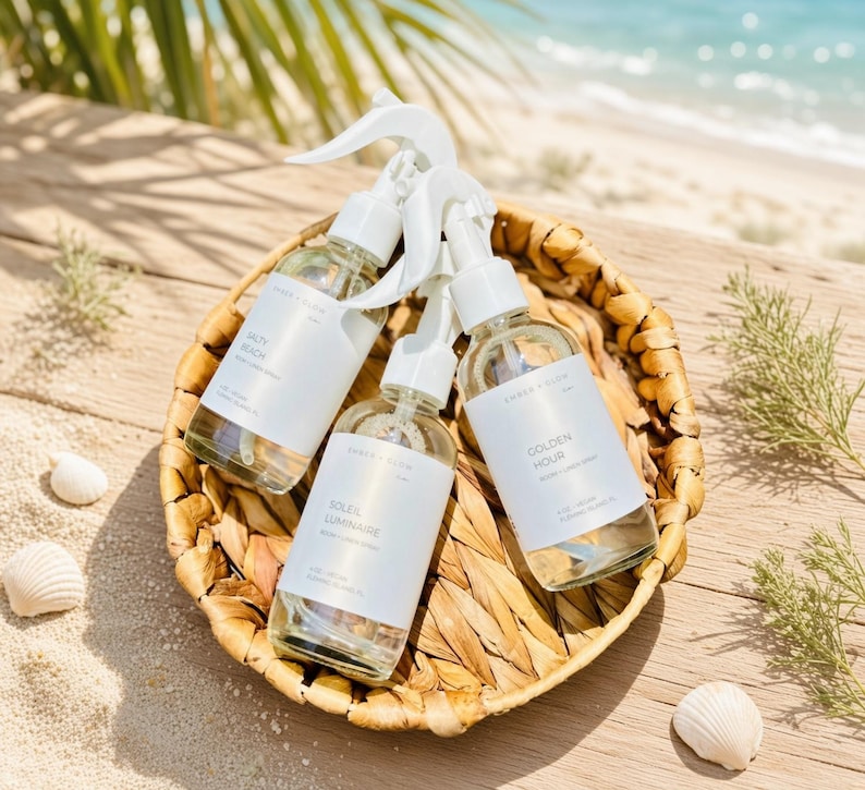 May include: Three clear glass spray bottles with white trigger nozzles and white labels, each containing a liquid. The labels feature the text Ember & Glow, with individual names Salty Beach, Soleil Luminaire, and Golden Hour printed on them.