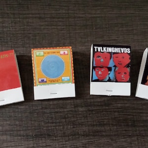 May include: Four matchbook covers featuring the band Talking Heads. The covers are red, yellow, blue, and black with different images of the band members.