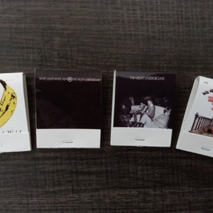 May include: Four matchbook covers featuring album art from The Velvet Underground. The covers feature a banana, a black and white photo, a black and white photo of a band, and a pink and white illustration of a bridge.