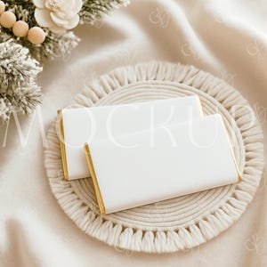 May include: Two white chocolate bars with gold foil edges rest on a macrame coaster. A sprig of frosted pine and a white flower are in the background, all on a cream-colored blanket.