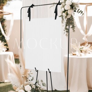 Peut inclure: Un panneau blanc avec des accents de ruban noir est affiché sur un chevalet noir. Le panneau est vierge et prêt pour un message. Le panneau est devant un fond blanc avec de la verdure et des fleurs blanches.